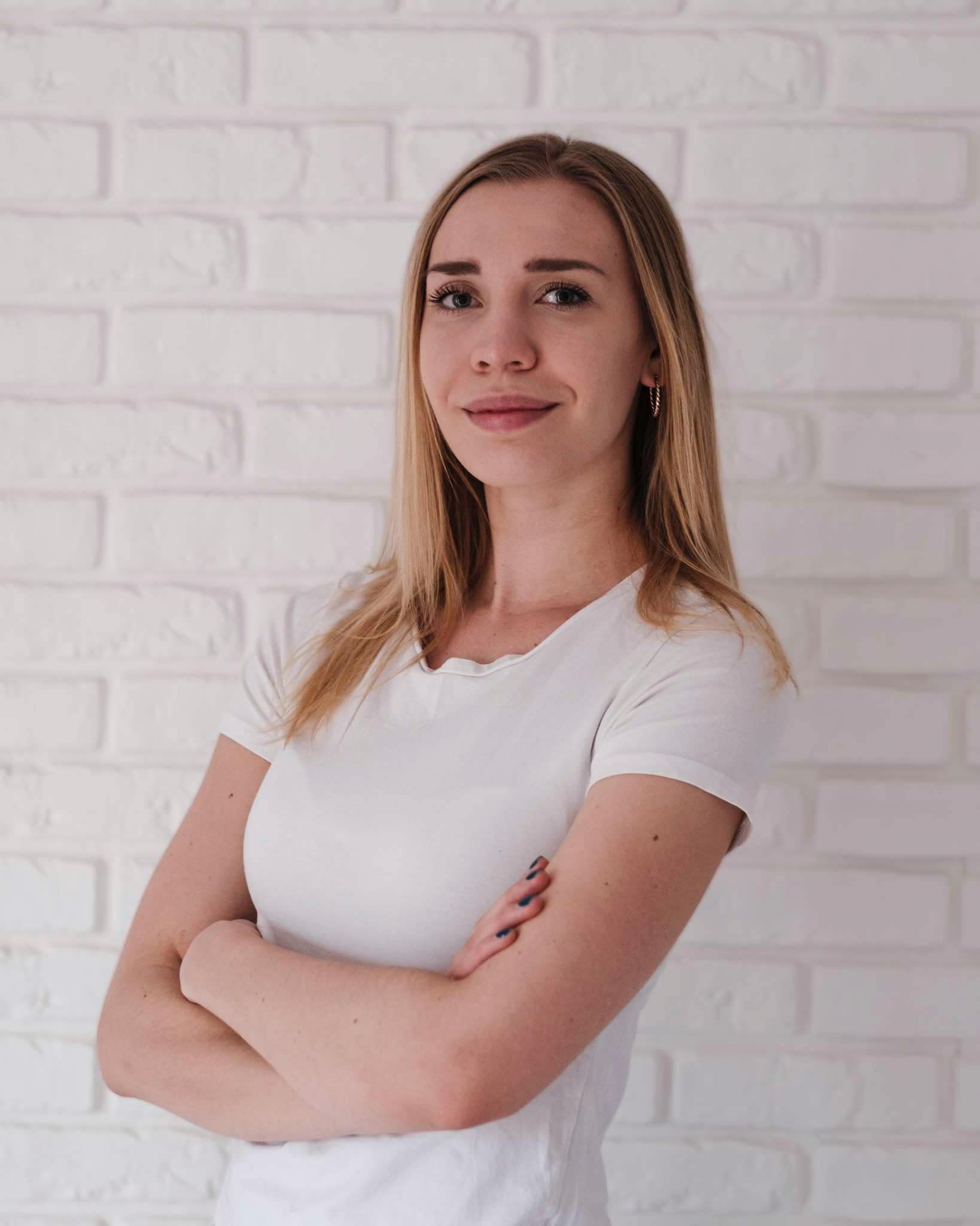 Obraz przedstawia Alę Buchlę, fizjoterapeutkę, młodą kobietę o blond włosach, ubraną w białą koszulkę, stojącą z założonymi rękoma na tle białej ceglanej ściany.
