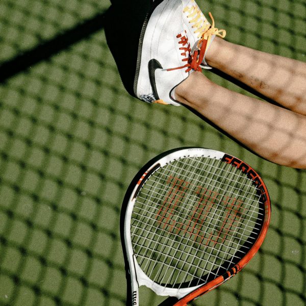 Obraz przedstawia fragment nóg w białych butach sportowych oraz rakietę tenisową leżące na zielonym korcie tenisowym z widocznym cieniem siatki.